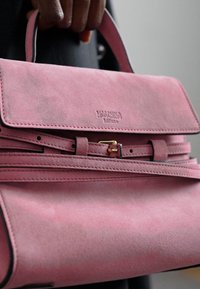 Rozā zamšādas rokassomā ar vāku, zeltīta aparatūra, un noņemams siksniņš. Priekšpusē redzams izspiedums ar zīmola logo.