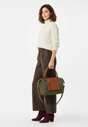 Frau in cremefarbenem Rollkragenpullover und brauner Hose mit weitem Bein, hält eine grüne und braune Handtasche, trägt bordeauxrote Stiefeletten und steht seitlich.