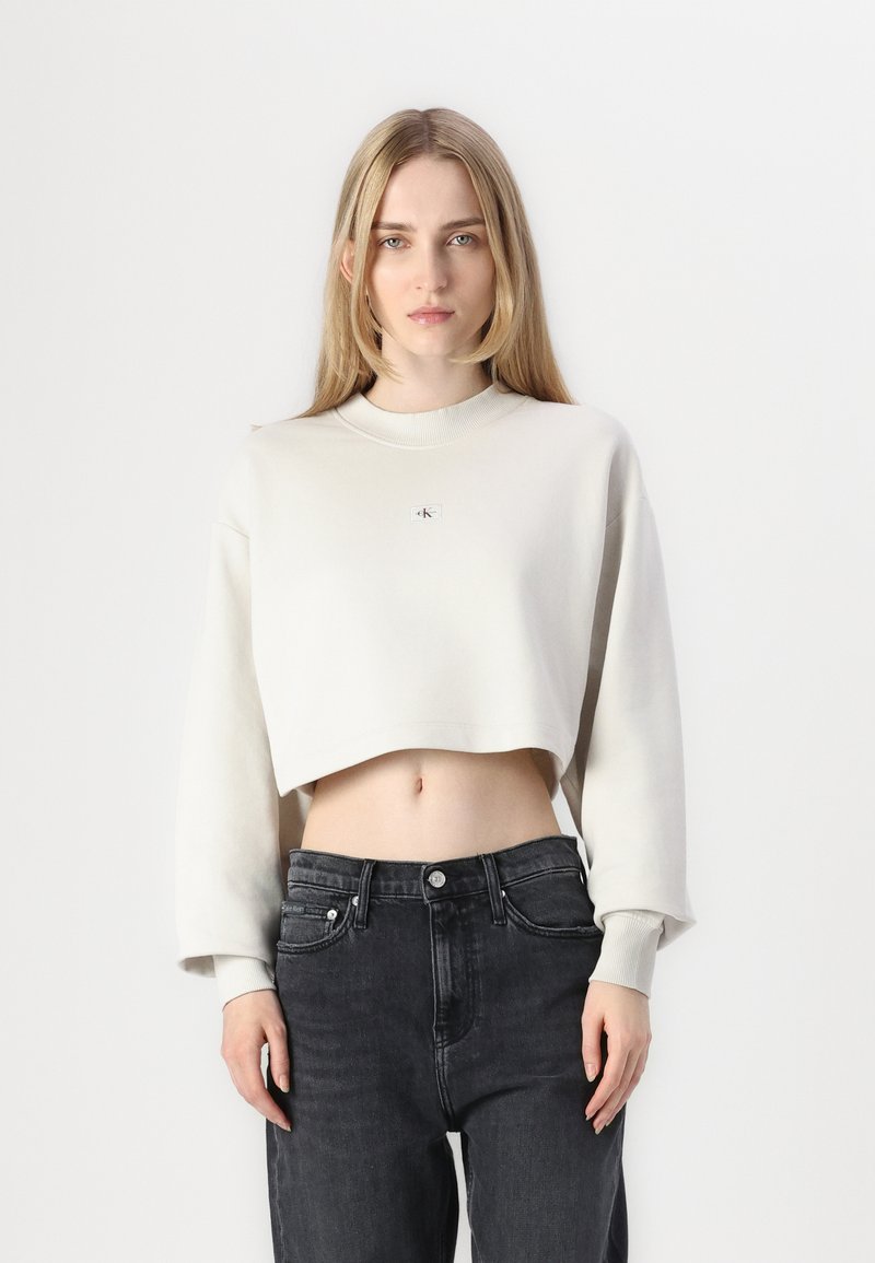 Cropped sweatshirt in light beige, featuring long sleeves and a round neck, with a small logo patch on the chest. Paired with dark jeans.