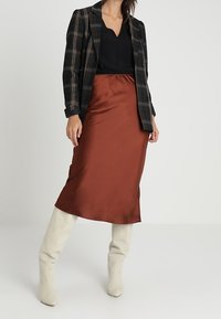 Brauner Satin-Midirock, schwarzes V-Ausschnitt-Top und karierter Blazer. Weiße kniehohe Stiefel runden das Outfit ab. Glatte Textur und taillierter Schnitt.