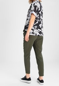 Kortärmad, svart skjorta med vitt blommönster, matchad med olivgröna avsmalnande byxor. Modellen bär svarta sneakers och står i profil.