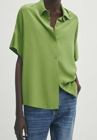 Chemise verte à manches courtes à boutons en tissu lisse, avec une coupe décontractée et un col pointu, assortie à un jean en denim bleu.