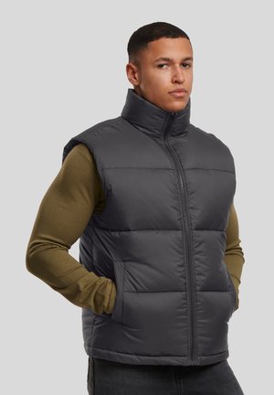Homme portant une veste matelassée sans manches gris foncé sur une chemise à manches longues vert olive, debout les mains dans les poches de la veste.