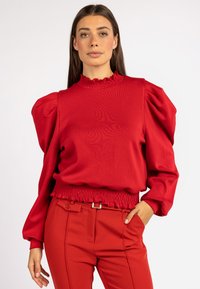 Roter Pullover mit Puffärmeln, Rüschenausschnitt und gerafftem Saum. Kombiniert mit roten, taillierten Hosen, die einen Gürtel mit einer metallischen Schnalle haben.