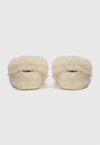 Crème-kleurige sloffen met een fluffy schapenwoltextuur, ronde vorm en zachte, gevoerde constructie; minimalistisch ontwerp zonder zichtbare hardware.