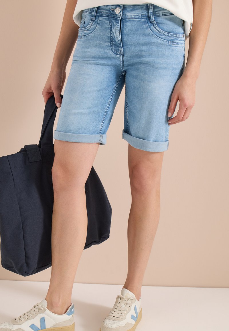 Short en jean bleu clair retroussé porté avec des baskets beige, tenant un grand sac cabas noir, sur un fond neutre.