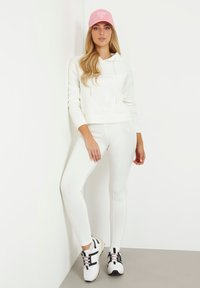 Sweatshirt à capuche blanc avec logo, associé à un legging blanc près du corps. Des baskets noir et blanc viennent compléter le look. Casquette rose en accent.