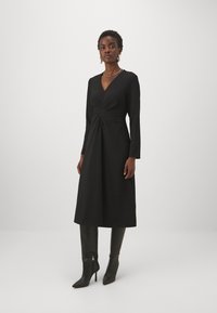 Abito midi nero con maniche lunghe e scollatura a V profonda, con dettaglio anteriore attorcigliato. La modella indossa stivali neri con tacco a spillo fino al ginocchio.