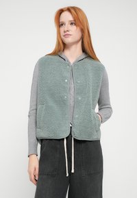 Colete de lã polar em verde claro com superfície texturizada, fecho de botões e bolsos laterais, usado sobre um casaco cinzento com fecho de correr e calças escuras.