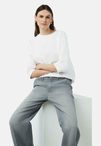 Witte sweatshirt met driekwartmouwen, gecombineerd met lichtgrijze wijde jeans. Model zit op een witte pedestal tegen een effen achtergrond.