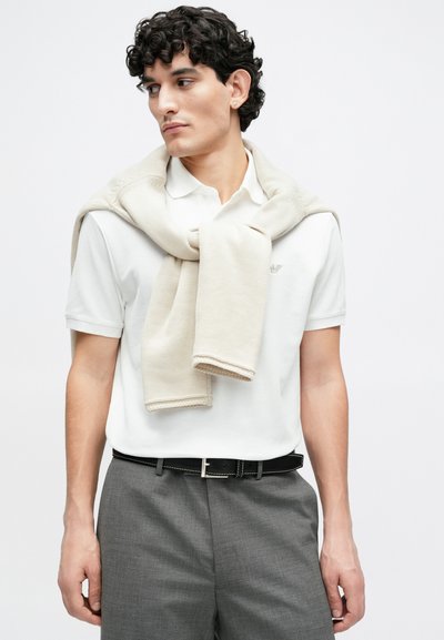 Biela polokošeľa, svetlo béžový sveter prehodený cez ramená, sivé nohavice a čierny opasok so striebornou prackou. Viditeľné kučeravé vlasy.