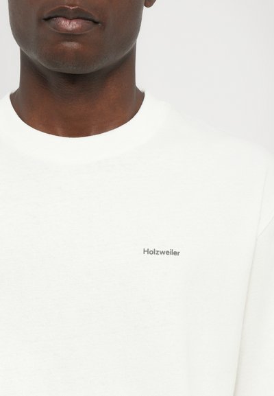 Sudadera blanca con un escote redondeado y una textura suave. Presenta un pequeño logo "Holzweiler" en gris en el lado izquierdo.