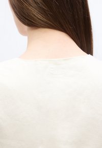 Πίσω όψη ενός ατόμου με ίσια καστανά μαλλιά που φοράει λευκό πουκάμισο, δείχνοντας το ανώτερο μέρος της πλάτης και την περιοχή του λαιμού.