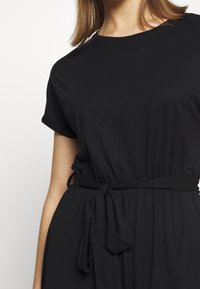 Femme portant une robe noire à manches courtes avec un col rond et une ceinture nouée à la taille, coupée au niveau des épaules et de la taille.