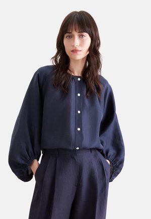 Vrouw met donker golvend haar draagt een marineblauwe blouse met knopen en pofmouwen en bijpassende hooggetailleerde broek, handen in de zakken.