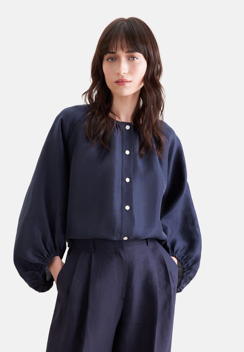 Femme aux cheveux bruns ondulés porte un chemisier bleu marine boutonné avec des manches bouffantes et un pantalon taille haute assorti, mains dans les poches.