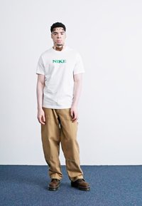 Hvid kortærmet Nike t-shirt med grøn logo, parret med beige posede bukser og mørkebrune sko, stående på et blåt tæppe.