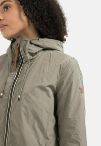 Lightweight, olive green jacket with a hood, featuring a zipper closure, drawstring accents, and a subtle logo patch on the sleeve.