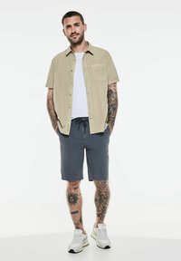 Chemise beige à manches courtes sur un T-shirt blanc, associée à un short gris foncé et des baskets blanches ; tatouages remarquables sur les bras.