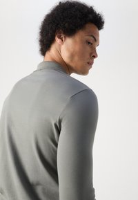 Lichtgrijze poloshirt met lange mouwen, een gestructureerde afwerking, een gespreide kraag en een aansluitende silhouet, gedragen door een model dat van de camera wegdraait.