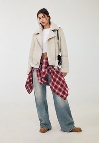Beige Schaffelljacke, weißes Croptop, rotkariertes Flanellhemd um die Taille gebunden, hellblaue weit geschnittene Jeans und beige Mokassins.