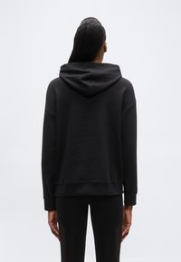 Άτομο με μακριά πλεξούδα, φορ ώντας μαύρο φούτερ με κουκούλα και μαύρα κολάν, όρθιο με την πλάτη γυρισμένη σε ένα απλό φωτεινό φόντο.