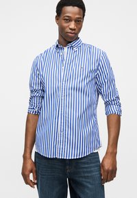 Μπλε και λευκό ριγέ πουκάμισο με κουμπιά, με μυτερό γιακά, γυρισμένα μανίκια και μικρό λογότυπο στο στήθος. Φορεμένο με σκουρόχρωμο τζιν.