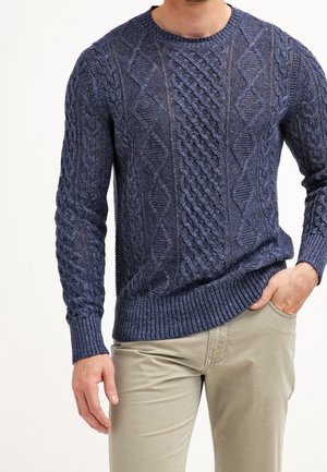 Mand iført en mørkeblå kabelstrikket sweater og beige bukser med den ene hånd i lommen mod en ensfarvet baggrund.