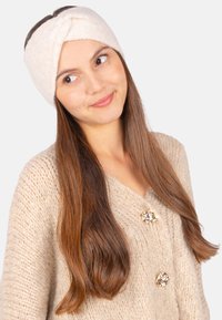 Un bandeau en tricot couleur crème avec un design torsadé, porté avec un cardigan épais beige à accents de boutons en or.