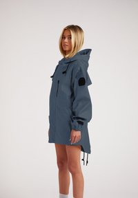 Chaqueta azul de gran tamaño, resistente al agua, con capucha, detalles en negro, puños elásticos y cordones ajustables. Cuenta con un bolsillo con cremallera.