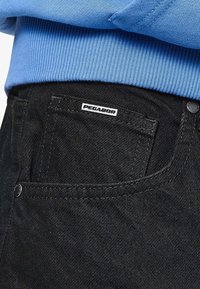 Vaqueros de mezclilla negra con un bolsillo trasero que presenta una etiqueta blanca "PEGADOR", acompañados de un suéter de punto azul claro.