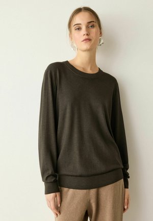 Ung kvinde med lyst hår iført en mørk langærmet sweater og beige bukser, står foran en ensartet lys baggrund.