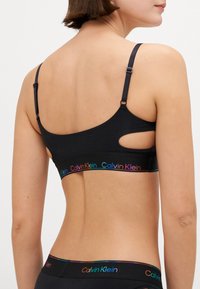 Svart sports-bh med justerbare stropper, utskjæringsdetaljer på siden, og et flerfarget Calvin Klein-logo bånd nederst.