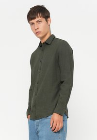 Camicia a maniche lunghe verde scuro in tessuto testurizzato, con frontale a bottoni e colletto, abbinata a jeans in denim blu chiaro.