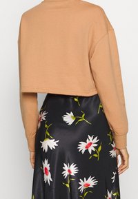 Avkortad, ljusbrun sweatshirt med lång ärm, som har en mjuk textur. Bärs över en marinblå kjol prydd med vita och röda blommönster.