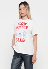 Jauna sieviete ar garām matiem, kas tērpusies baltā T-kreklā ar sarkanu uzrakstu "Slow Coffee Club" un zilām kafijas tematiskām ilustrācijām, apvienojot ar tumšām biksēm.