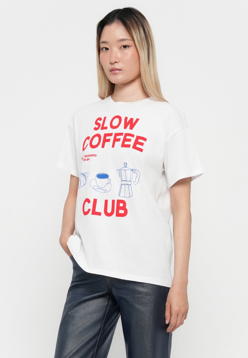 Giovane donna con i capelli lunghi, che indossa una maglietta bianca con la scritta rossa "Slow Coffee Club" e illustrazioni a tema caffè in blu, abbinata a pantaloni scuri.