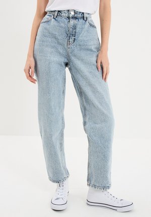 Person trägt hellblaue hoch taillierte Jeans, weiße Sneaker und ein in die Hose gestecktes weißes Hemd und steht vor weißem Hintergrund.