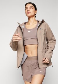 Veste imperméable marron clair avec capuche, poches zippées et accents réfléchissants, associée à un soutien-gorge de sport et un short assortis.