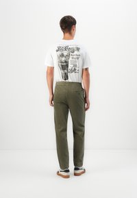 T-shirt bianca a maniche corte con grafica vintage nera di Pepsi sulla schiena, abbinata a pantaloni a righe verdi e sneakers bianche con dettagli neri.