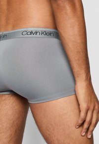 Graue Baumwollboxershorts mit einem logoed Bund, der eine glatte Textur und eine taillierte Passform aufweist, die einen Teil des Oberschenkels und der Haut zeigt.