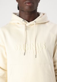 Hoodie beige chiaro con design a pullover, lacci con punte metalliche e logo "ORIGINALS" in rilievo sul petto. Tessuto dalla consistenza morbida.