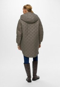 Quilted, olivengrøn lang frakke med hætte, der har en afslappet pasform og ribbede manchetter. Båret med mørke jeans og høje brune støvler.
