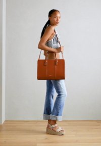Sac fourre-tout en cuir marron avec poignées doubles, surface lisse et design structuré. Porté avec un jean évasé bleu clair et des sandales à plateforme beiges.