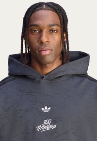 Musta huppari, jossa on tekstiilitason maastokuvio, valkoinen Adidas-logo ja valkoiset "100 Thieves" -tekstit. Hupparissa on kiristettävä huppu.