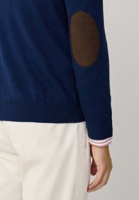 Suéter azul marino con un acabado texturizado y parches de cuero marrón en los codos. Presenta puños y dobladillo acanalados, superpuesto a una camisa a rayas.