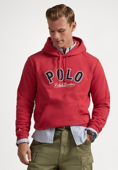 Muškarac u crvenoj Polo Ralph Lauren majici s kapuljačom preko plave prugaste košulje i zelenih cargo hlača, gleda dolje s rukama u džepovima.