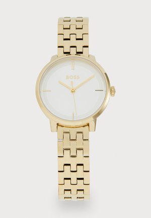 Montre en métal doré avec un cadran blanc, présentant un design simple, des index horaires et un bracelet à maillons. Nom de la marque affiché en évidence.