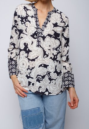 Kvinde iført langærmet bluse med sort-hvide blomster- og cirkelmønstre, kombineret med lyseblå jeans med denim lapper.