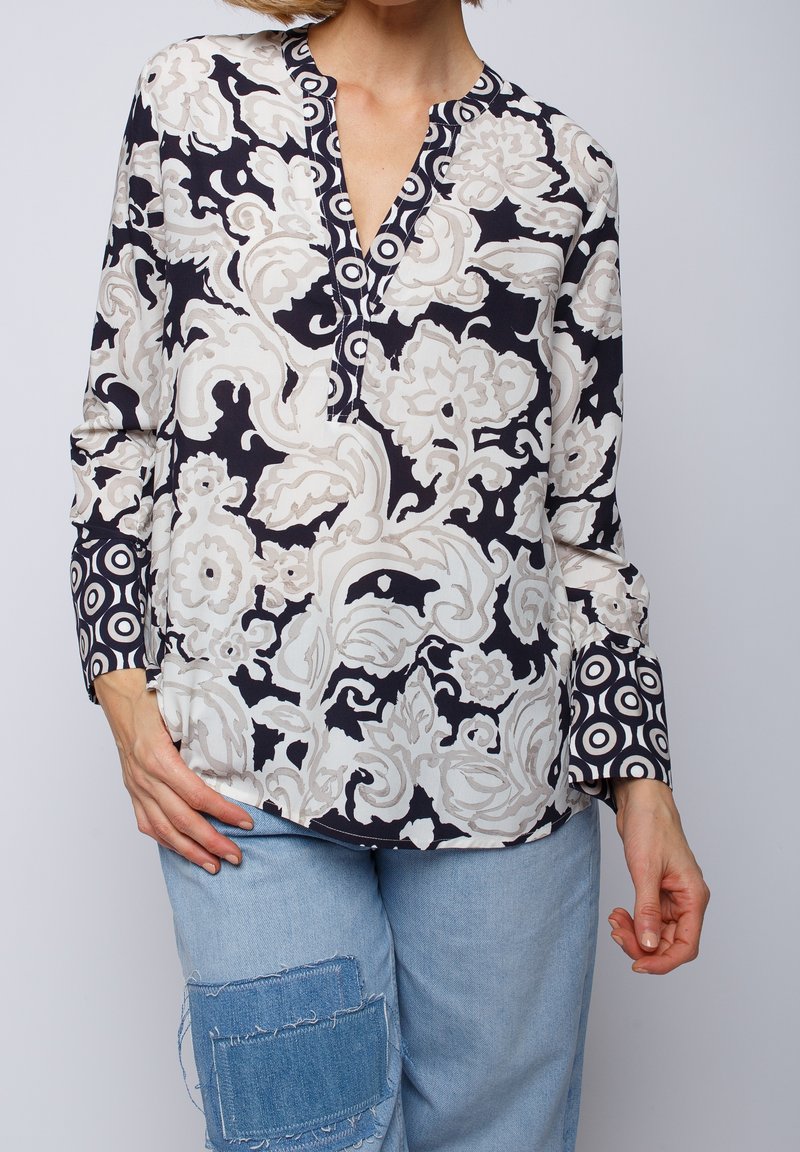 Femme portant un chemisier à manches longues avec des motifs floraux et circulaires noir et blanc, associé à un jean bleu clair avec patchwork en denim.